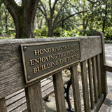 Wooden bench with a bronze plaque reading 'Honoring the past, Enjoying the present, Building the future' in a park setting.