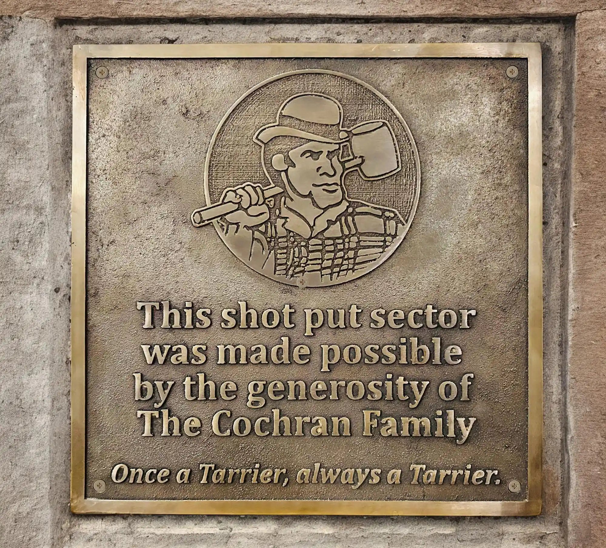Bronze plaque with a logo of a person holding an axe and engraved text on a stone wall.