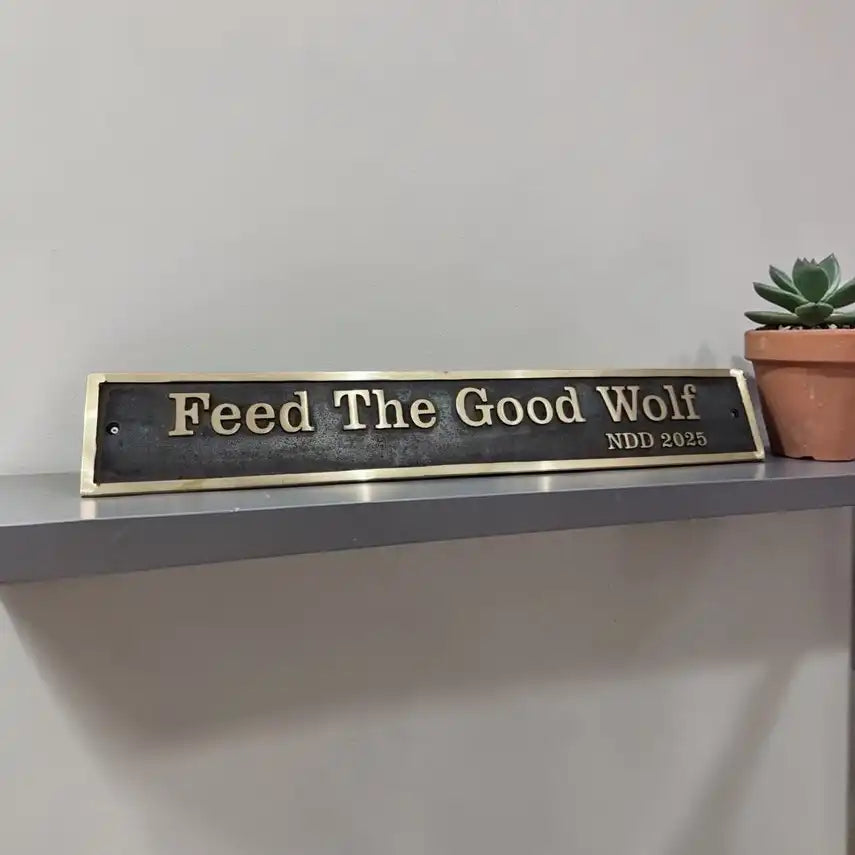 Bronze name sign with 'Feed The Good Wolf' text on a shelf with a plant in the background