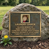 Bronze Memorial plaque for Pamie Cameron on a rock with a yellow flower in front, set against a natural background.