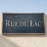 Bronze Street sign reading 'RUE DU LAC' on a textured wall with a clear blue sky.