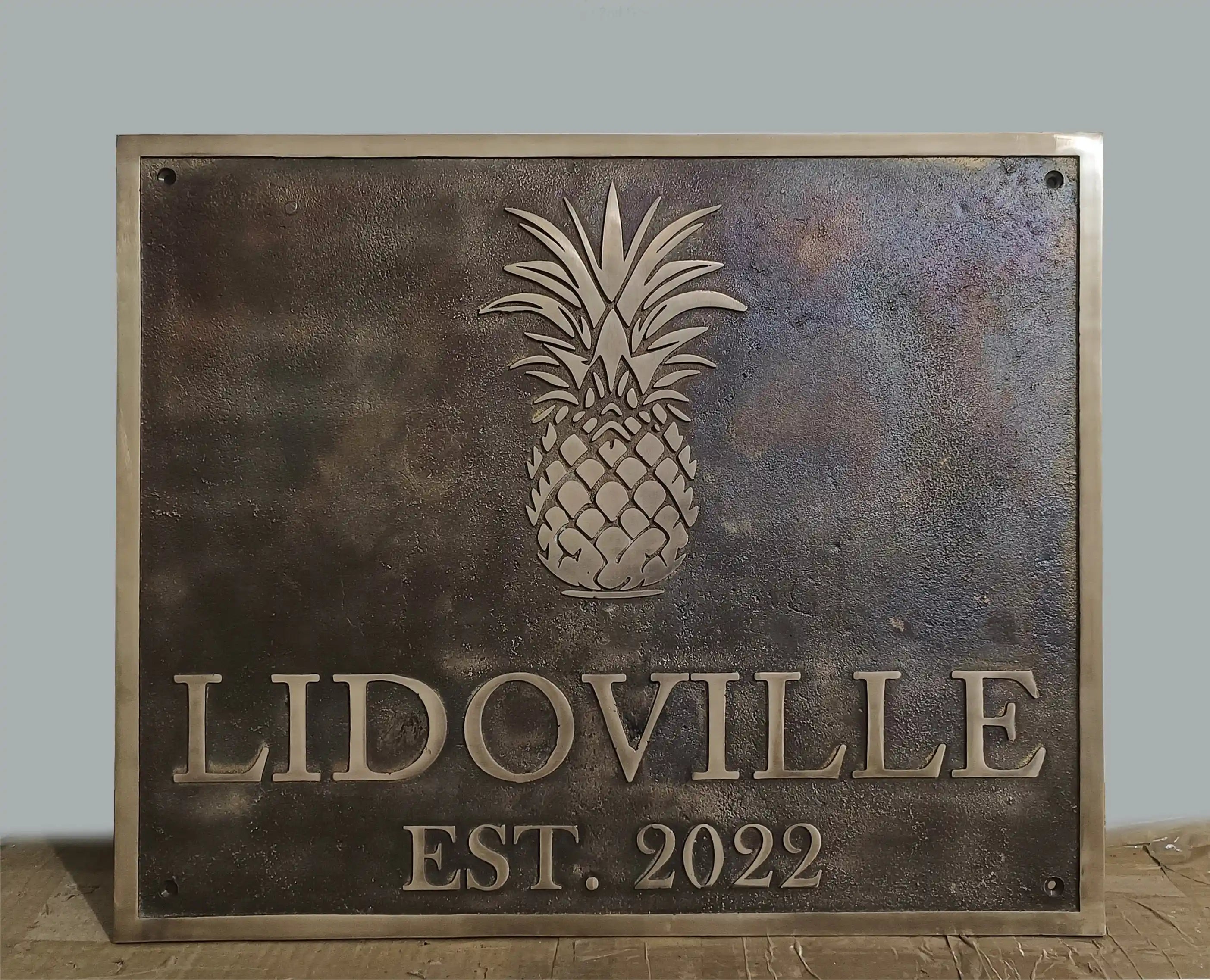 Metal sign with pineapple design and 'Lidoville Est. 2022' text on a wooden surface.