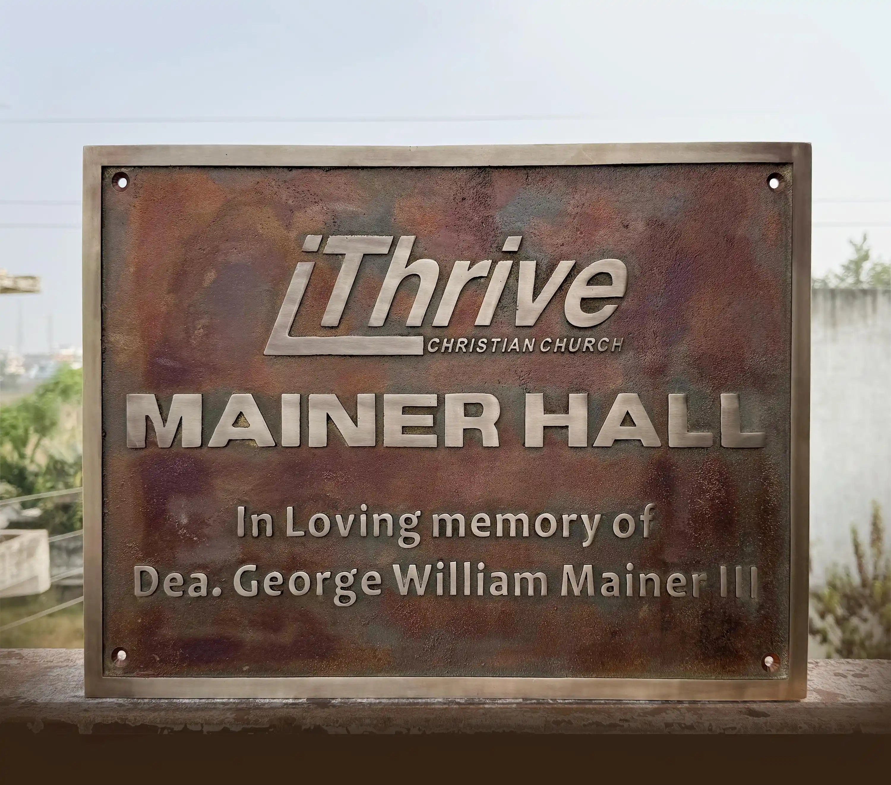 Metal plaque with 'iThrive Christian Church' and 'Mainer Hall' engraved on a blurred natural background