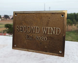Bronze plaque with 'SECOND WIND Est. 2020' text on a white surface with a blurred outdoor background.