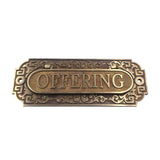 An ornate rectangular bronze metal sign with the word 'OFFERING' in raised capital letters inside an oval center, surrounded by decorative scrollwork and Greek key corner details, shown on a plain white background.