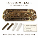 A custom brass plaque with decorative scrollwork and the placeholder text 'YOUR TEXT', shown with two matching screws and two white wall anchors on a white background. Text above and below reads 'CUSTOM TEXT' and highlights that the product is solid cast brass with an antique finish and includes matching screws and wall anchors.