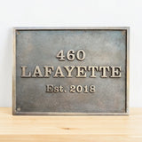 Bronze metal plaque with engraved text '460 Lafayette Est. 2018' on a wooden surface.