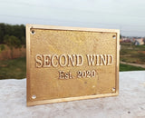 Gold plaque with 'SECOND WIND Est. 2020' text on a stone surface with a blurred natural background
