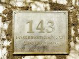 Metallic house number plaque with '143 Preservation Drive Love Lives Here' on a textured wall.
