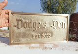 Brass sign with 'Dodge's Den EST. 2022' on a stone surface with a blurred outdoor background.
