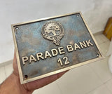 Metal plaque with 'Parade Bank 12' text held by a hand against a light-colored wall.