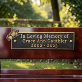 A close-up of a memorial plaque mounted on a dark wooden bench in an outdoor park setting. The plaque is black with gold lettering that reads 'In Loving Memory of Grace Ann Gauthier 2002 - 2023' and features a gold butterfly emblem on the left.