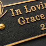 Close-up of a black textured plaque with raised gold lettering reading 'In Loving Grace' and a gold border, shown on a dark background.