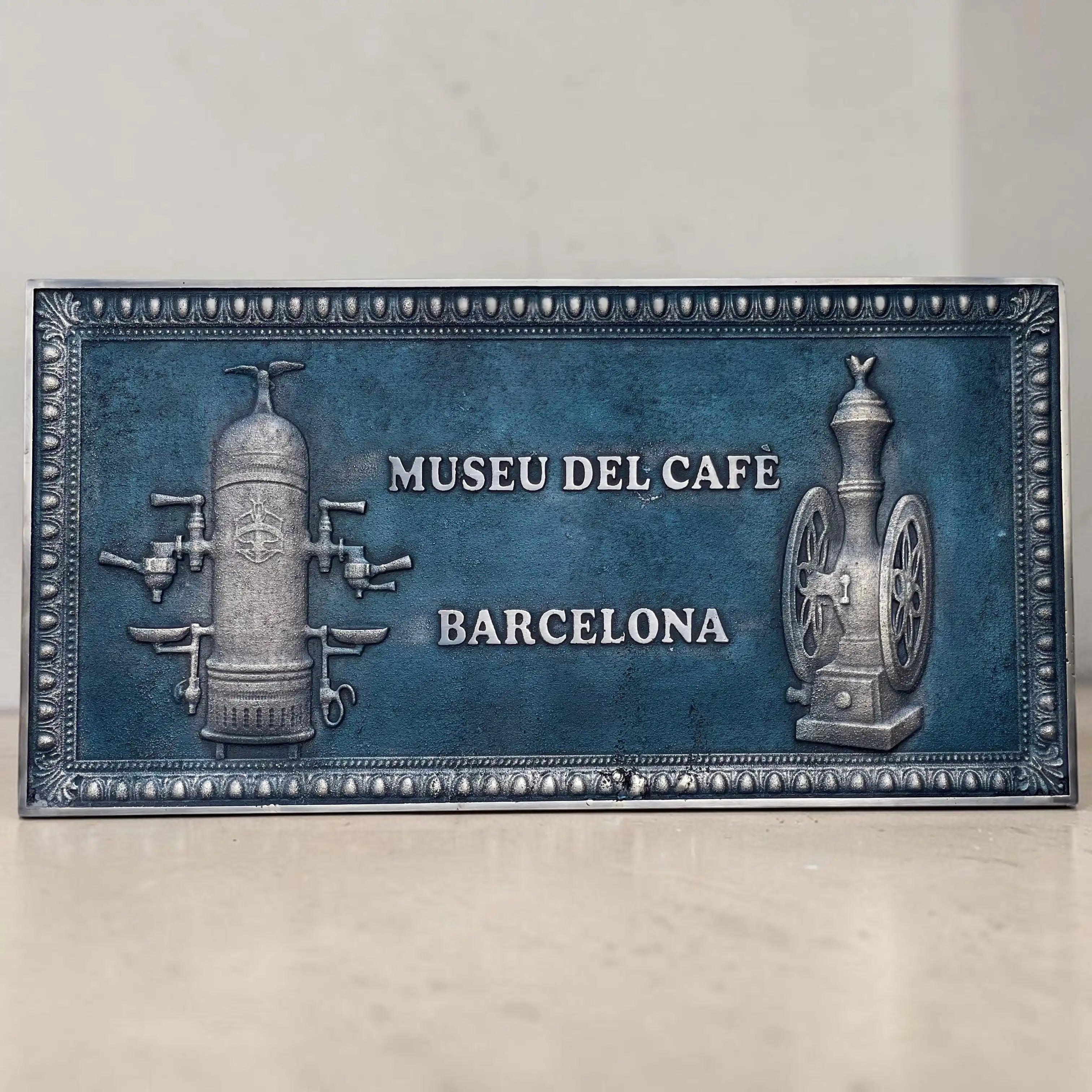 A rectangular decorative plaque featuring a blue textured background with embossed vintage coffee equipment on both sides and the text 'MUSEU DEL CAFE BARCELONA' in the center, photographed against a light beige wall and surface.