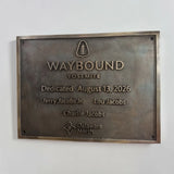 A rectangular bronze Waybound Yosemite memorial plaque mounted on a white wall. The plaque features raised text including 'WAYBOUND', 'Yosemite', the tree logo, the dedication date August 13, 2026, and names Jerry Jacobs Jr.