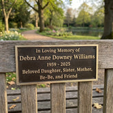 Memorial plaque on a bench in a park with a garden and trees in the background.
