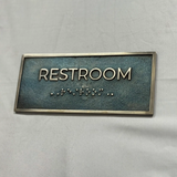 Rectangular sign with 'RESTROOM' text and braille on a gray background