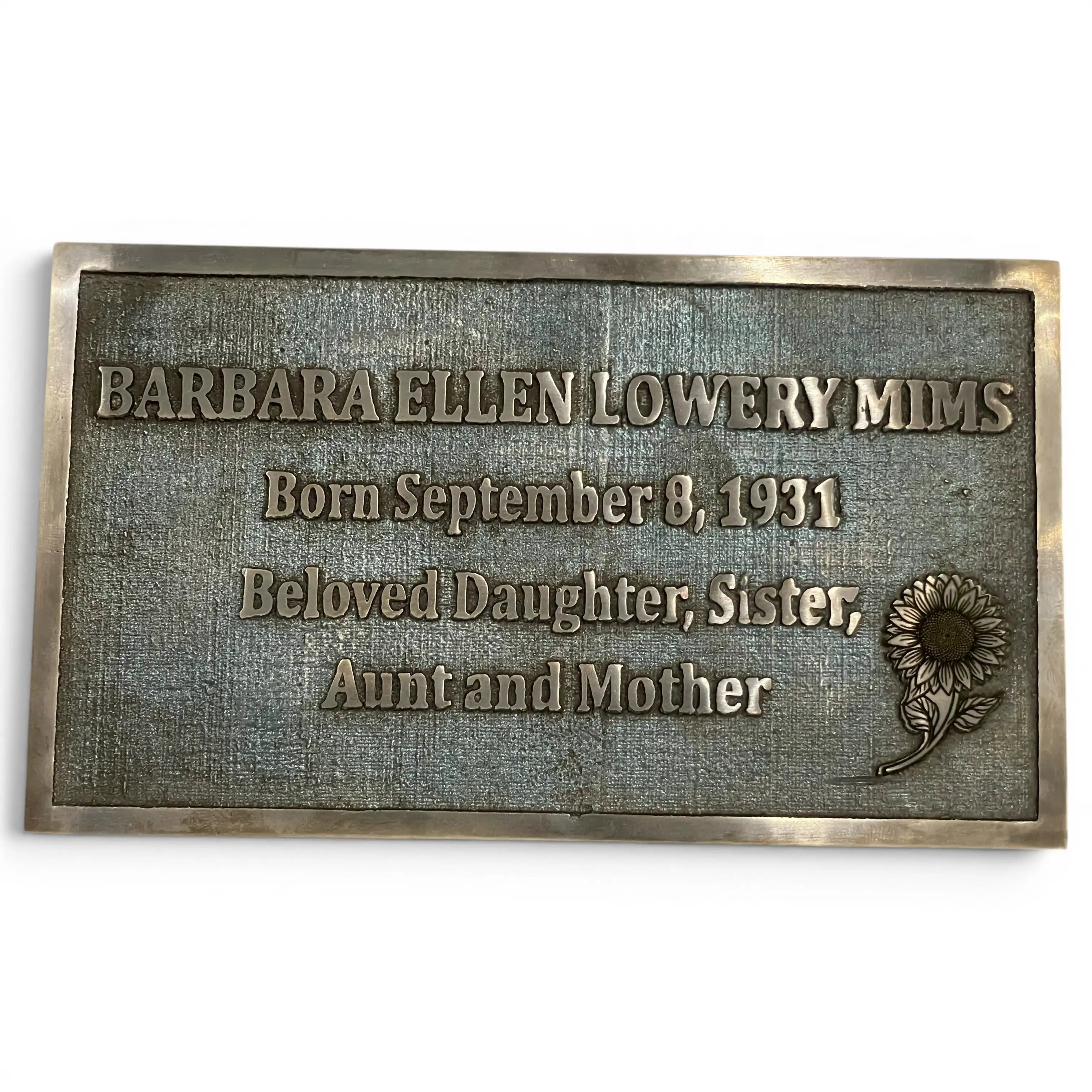 A rectangular bronze memorial plaque with engraved text reading 'BARBARA ELLEN LOWERY MIMS' and 'Born September 8, 1931 Beloved Daughter, Sister, Aunt and Mother,' plus an embossed sunflower design, shown on a plain white background.
