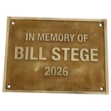 A rectangular bronze-toned metal memorial plaque with raised lettering that reads 'IN MEMORY OF BILL STEGE 2026' on a plain white background.
