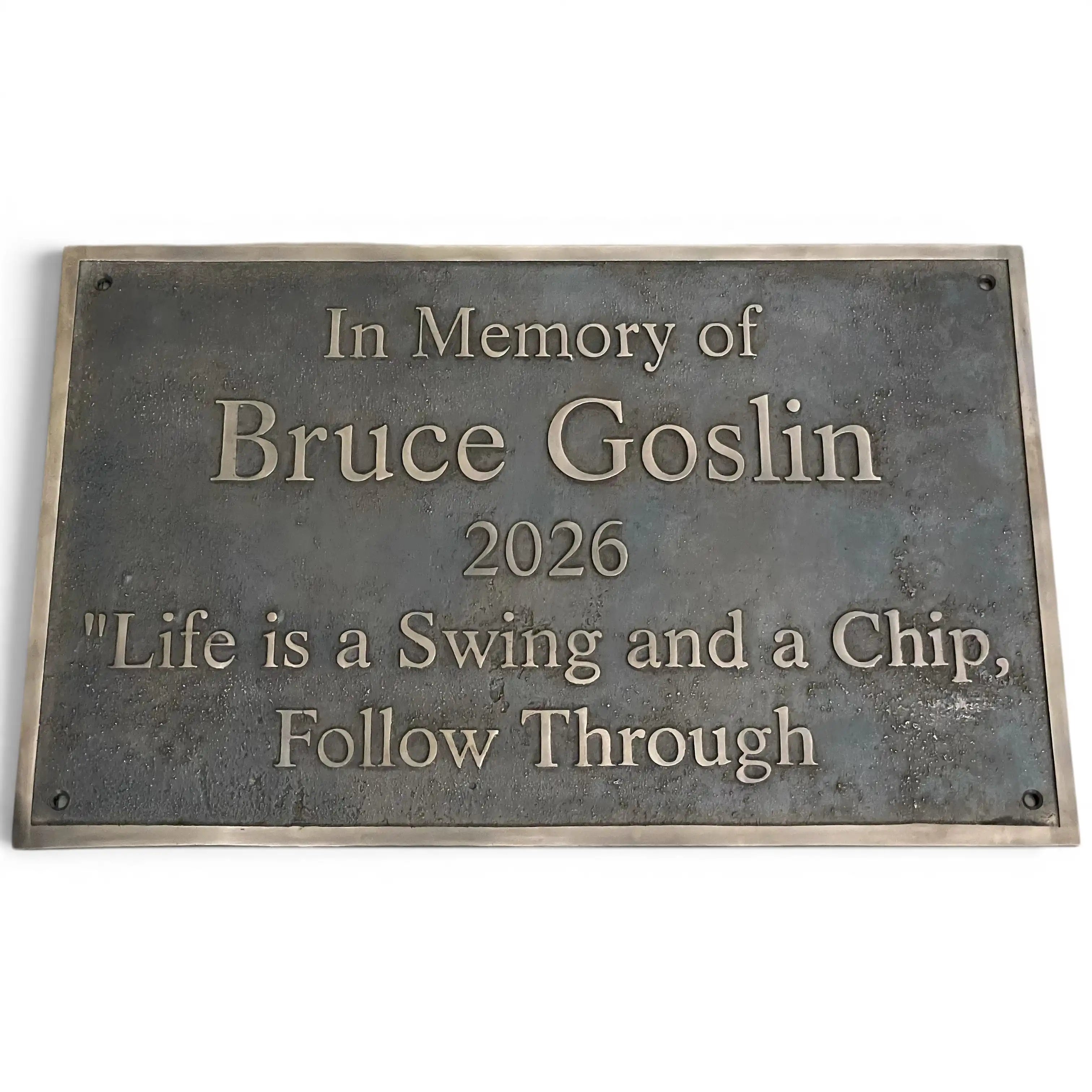 A rectangular bronze memorial plaque with raised lettering that reads 'In Memory of Bruce Goslin 2026 "Life is a Swing and a Chip, Follow Through"' on a plain white background.