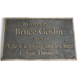 A rectangular bronze memorial plaque with raised lettering that reads 'In Memory of Bruce Goslin 2026 "Life is a Swing and a Chip, Follow Through"' on a plain white background.