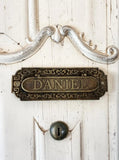 Decorative metal mail slot with 'DANIEL' on a white wooden door.