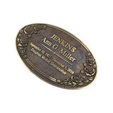 An oval bronze-toned metal keepsake plaque engraved with the name 'JENKINS Ann C. Miller' and dates, decorated with embossed floral rose designs around the border, shown on a plain white background.