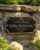 A personalized black memorial plaque with gold lettering mounted on a rustic stone wall, surrounded by green foliage and small white flowers. The plaque reads 'In Loving Memory John Anderson 1948-2025'.