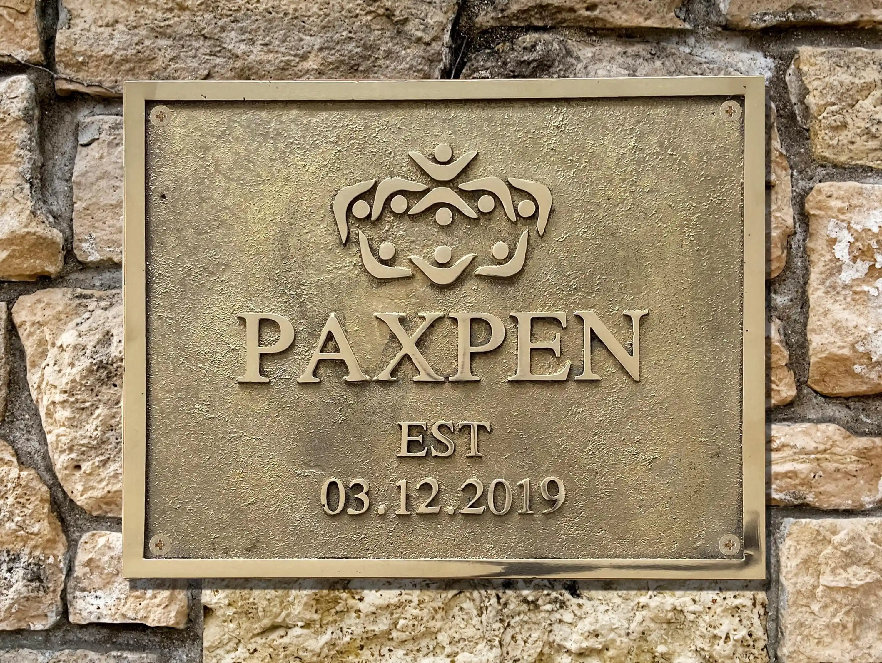 Bronze plaque with 'PAXPEN' logo and establishment date on a stone wall