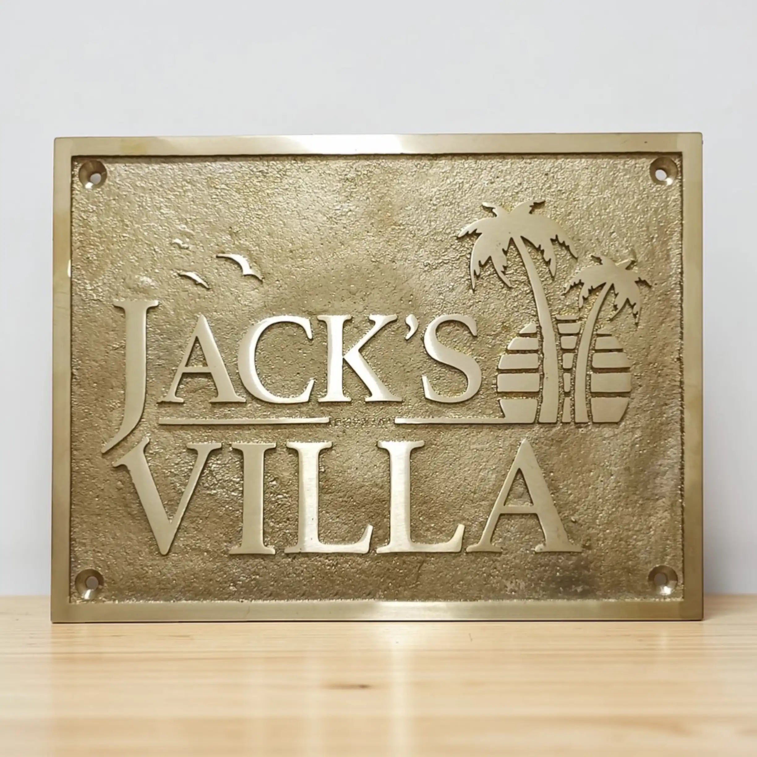 Brass finish Bronze plaque with 'Jack's Villa' text and palm tree design on a wooden surface.