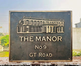 Metal sign with architectural illustration and text 'The Manor No 9 GT Road' against a blurred outdoor background.