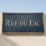 Bronze Street sign reading 'RUE DU LAC' on a textured wall with a clear blue sky.