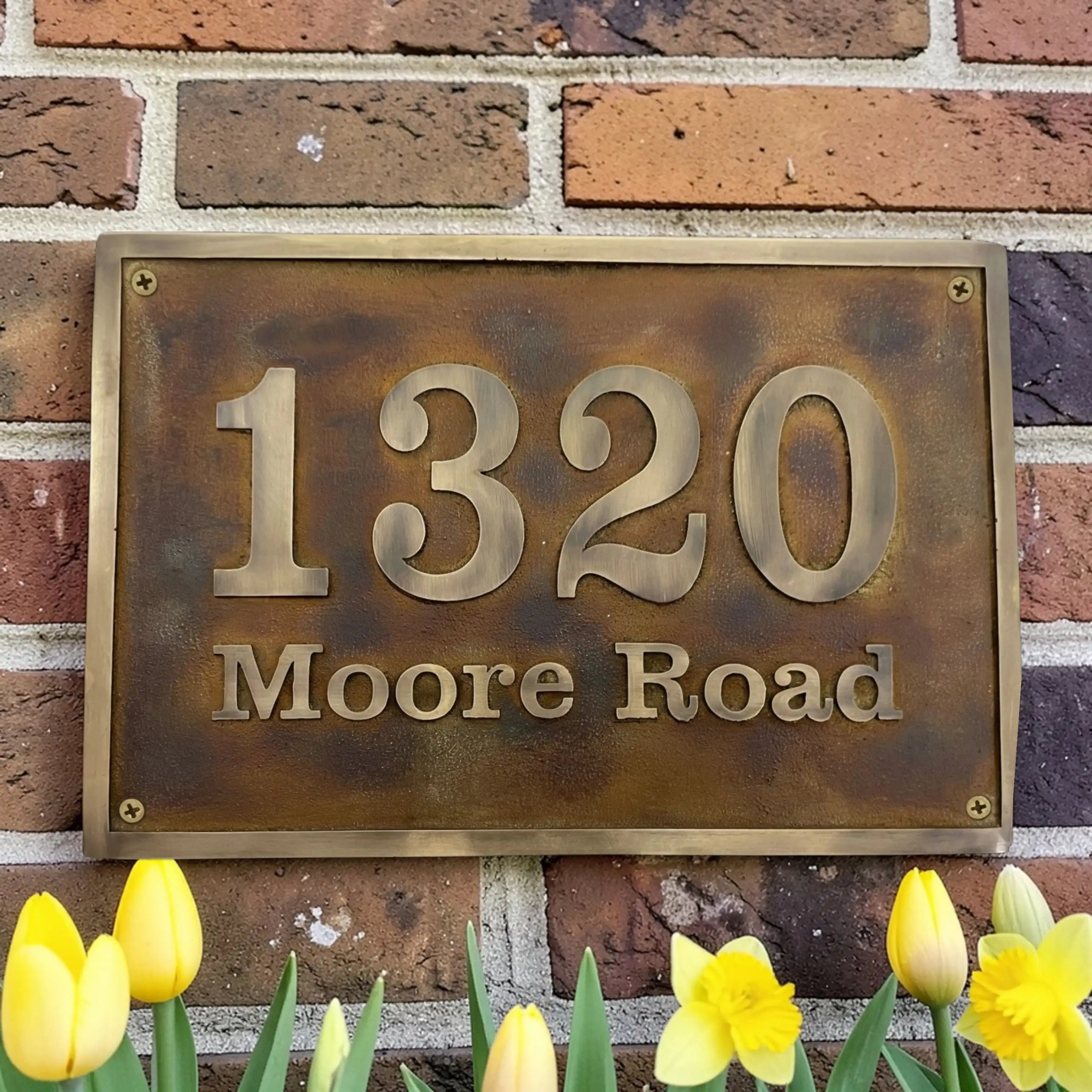 Brass house number plaque with '1320 Moore Road' on a brick wall with flowers in the foreground.