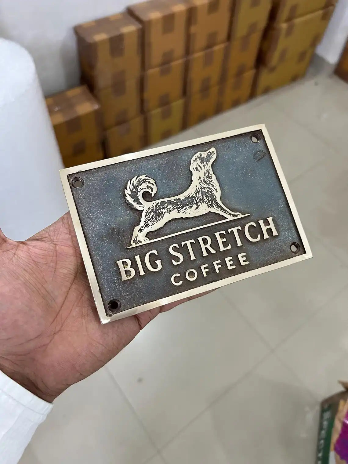 Hand holding a 'Big Stretch Coffee' sign with a dog logo against a background of stacked boxes.