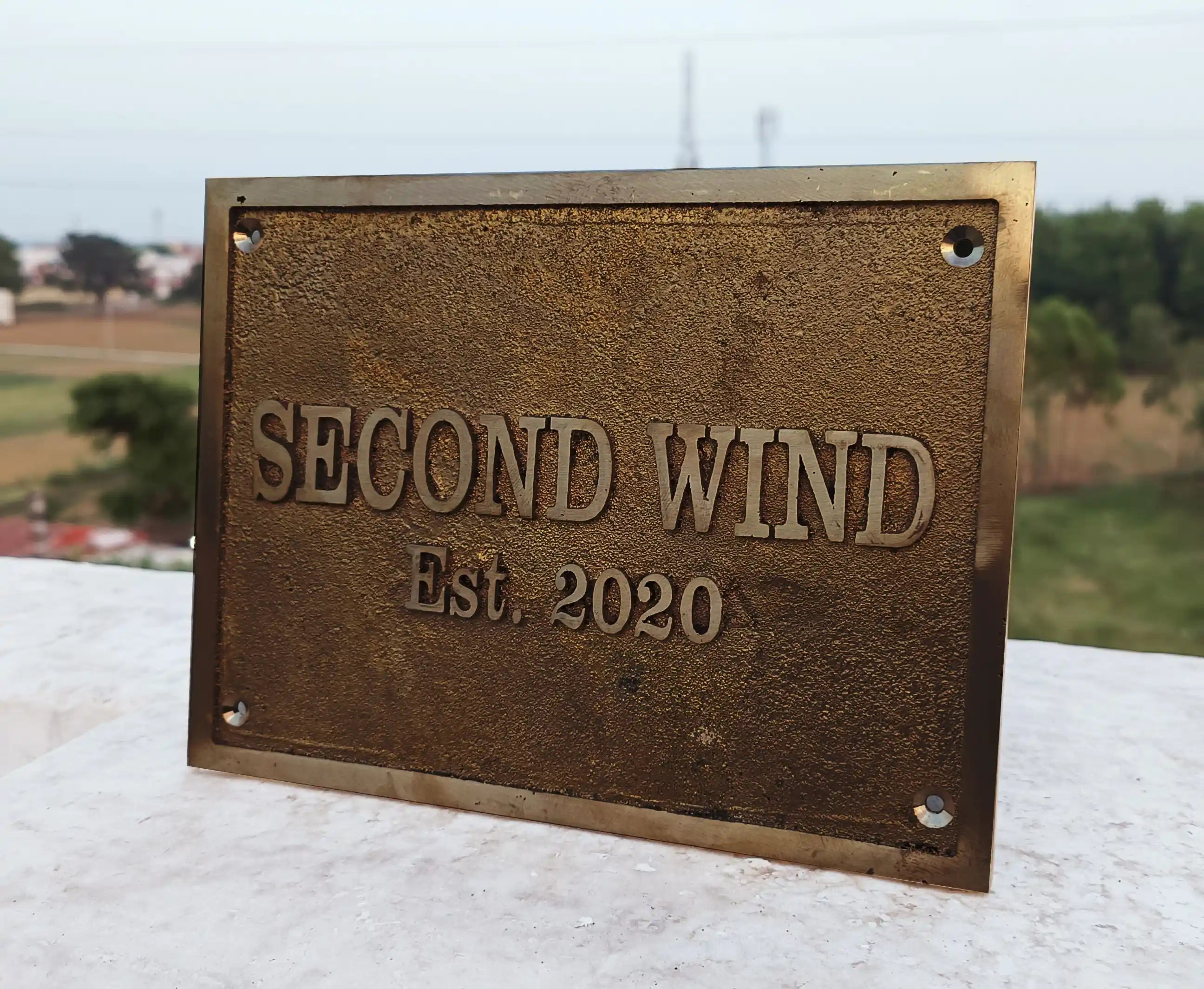 Bronze plaque with 'SECOND WIND Est. 2020' text on a white surface with a blurred outdoor background.