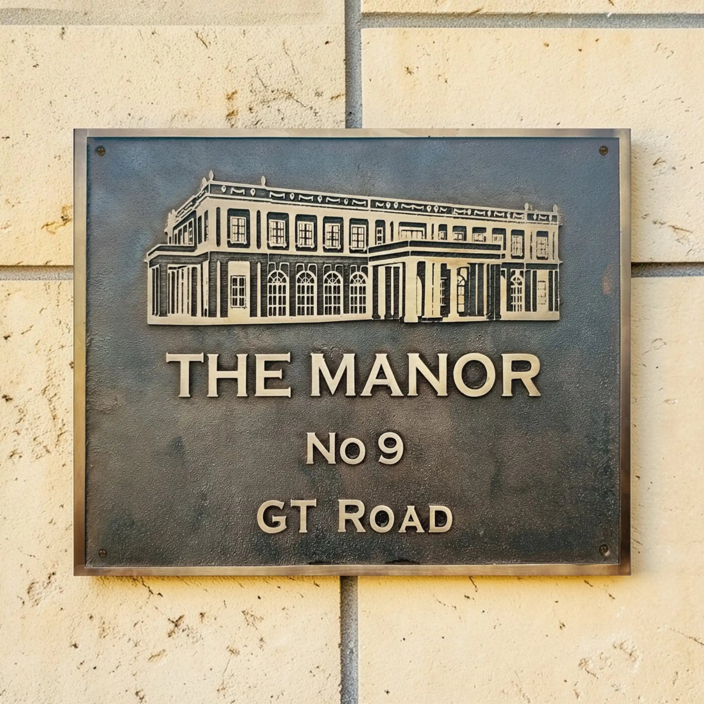 Bronze address sign with architectural illustration and text 'The Manor No 9 GT Road' on a stone wall.