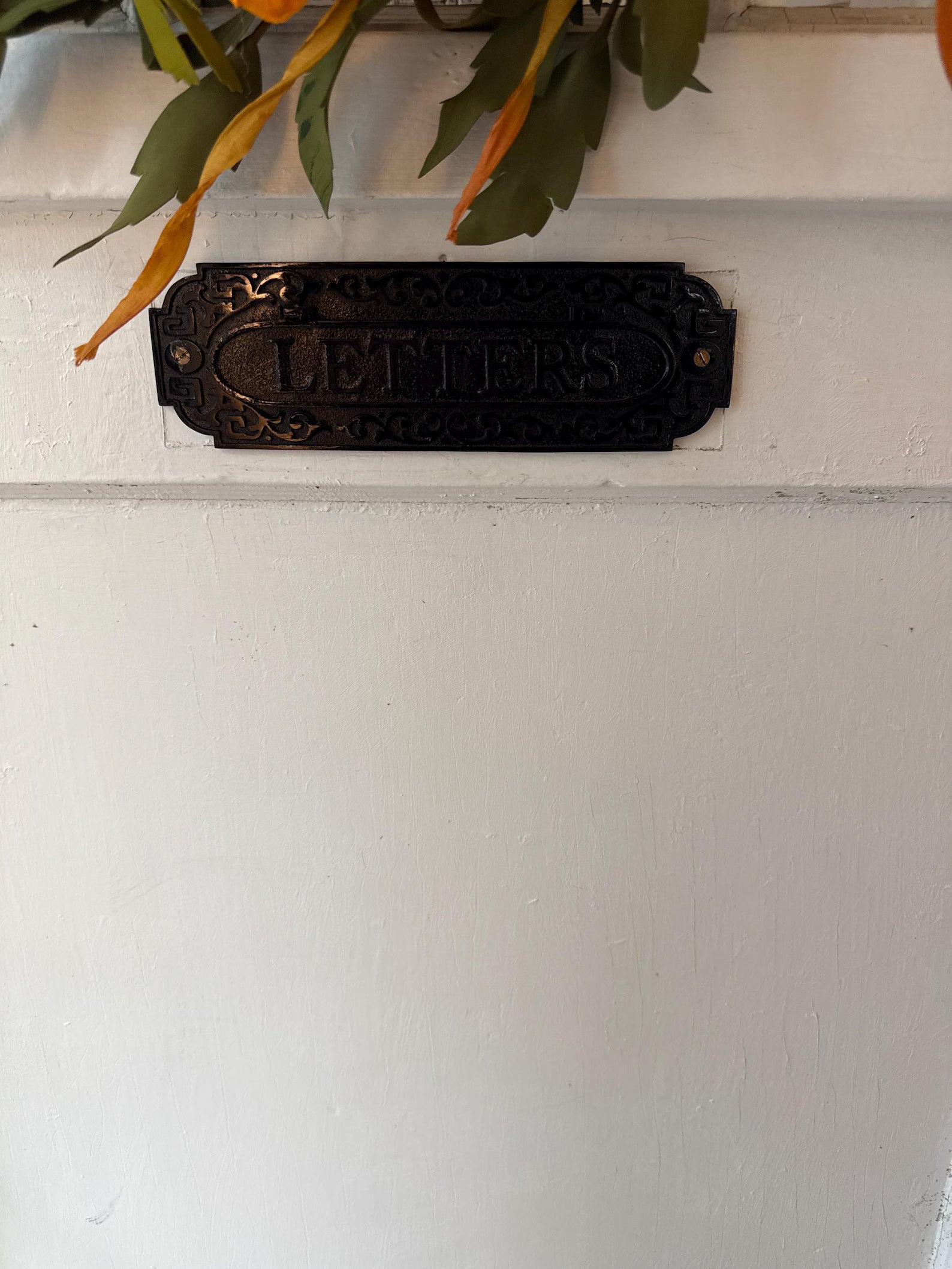 Decorative metal letter box sign on a white wall with green leaves above.