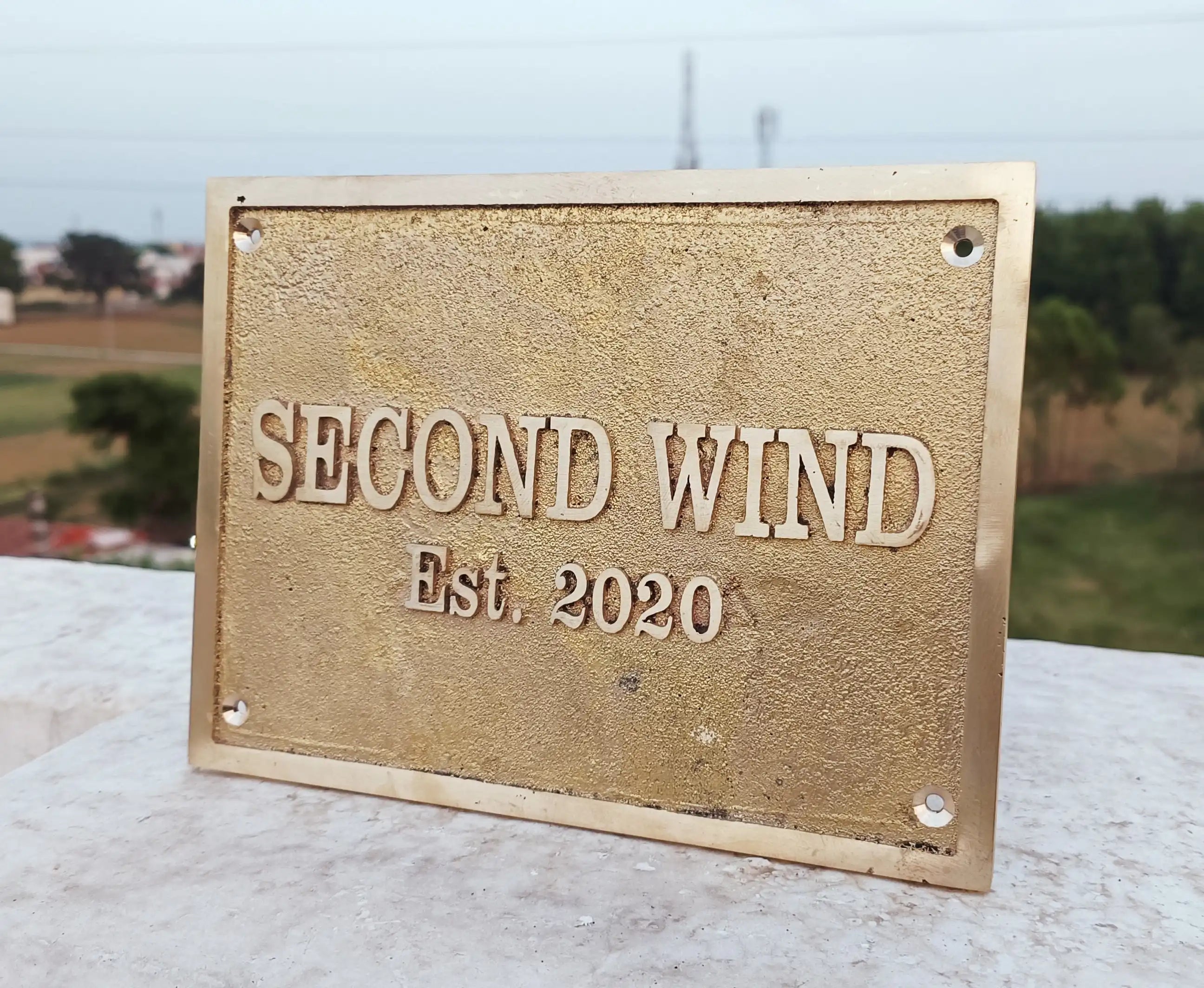 Brass plaque with 'SECOND WIND Est. 2020' text on a stone surface with a blurred outdoor background.