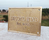 Brass plaque with 'SECOND WIND Est. 2020' text on a stone surface with a blurred outdoor background.