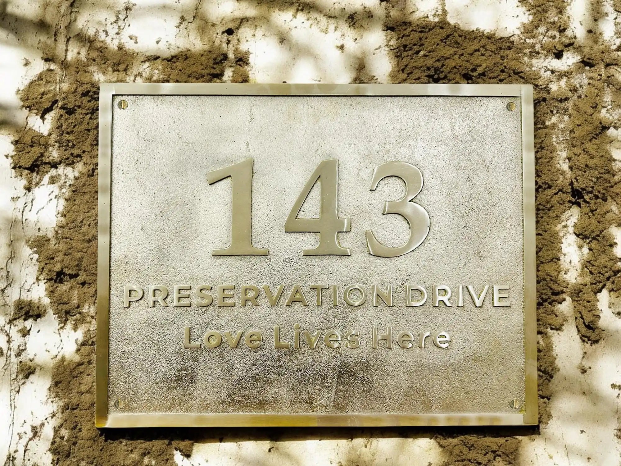 Metallic house number plaque with '143 Preservation Drive Love Lives Here' on a textured wall.