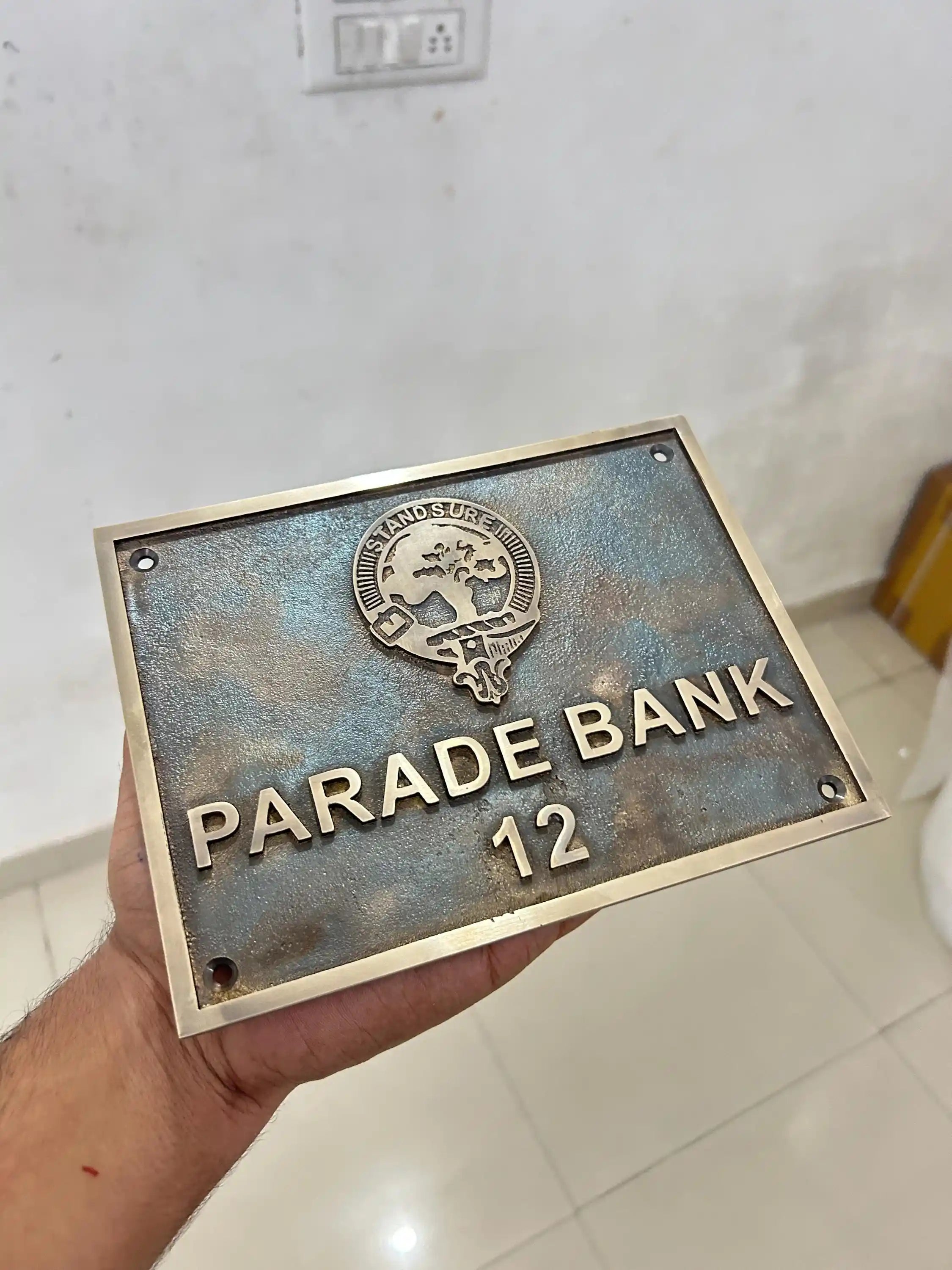 Metal plaque with 'Parade Bank 12' text held by a hand against a light-colored wall.