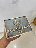 Metal plaque with 'Parade Bank 12' text held by a hand against a light-colored wall.