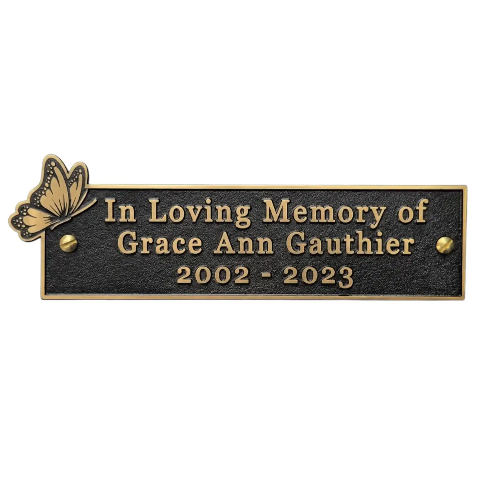 A rectangular black memorial plaque with gold-toned lettering that reads 'In Loving Memory of Grace Ann Gauthier 2002 - 2023,' plus a small butterfly motif on the left and gold-toned border details.