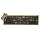 A rectangular black memorial plaque with gold-toned lettering that reads 'In Loving Memory of Grace Ann Gauthier 2002 - 2023,' plus a small butterfly motif on the left and gold-toned border details.