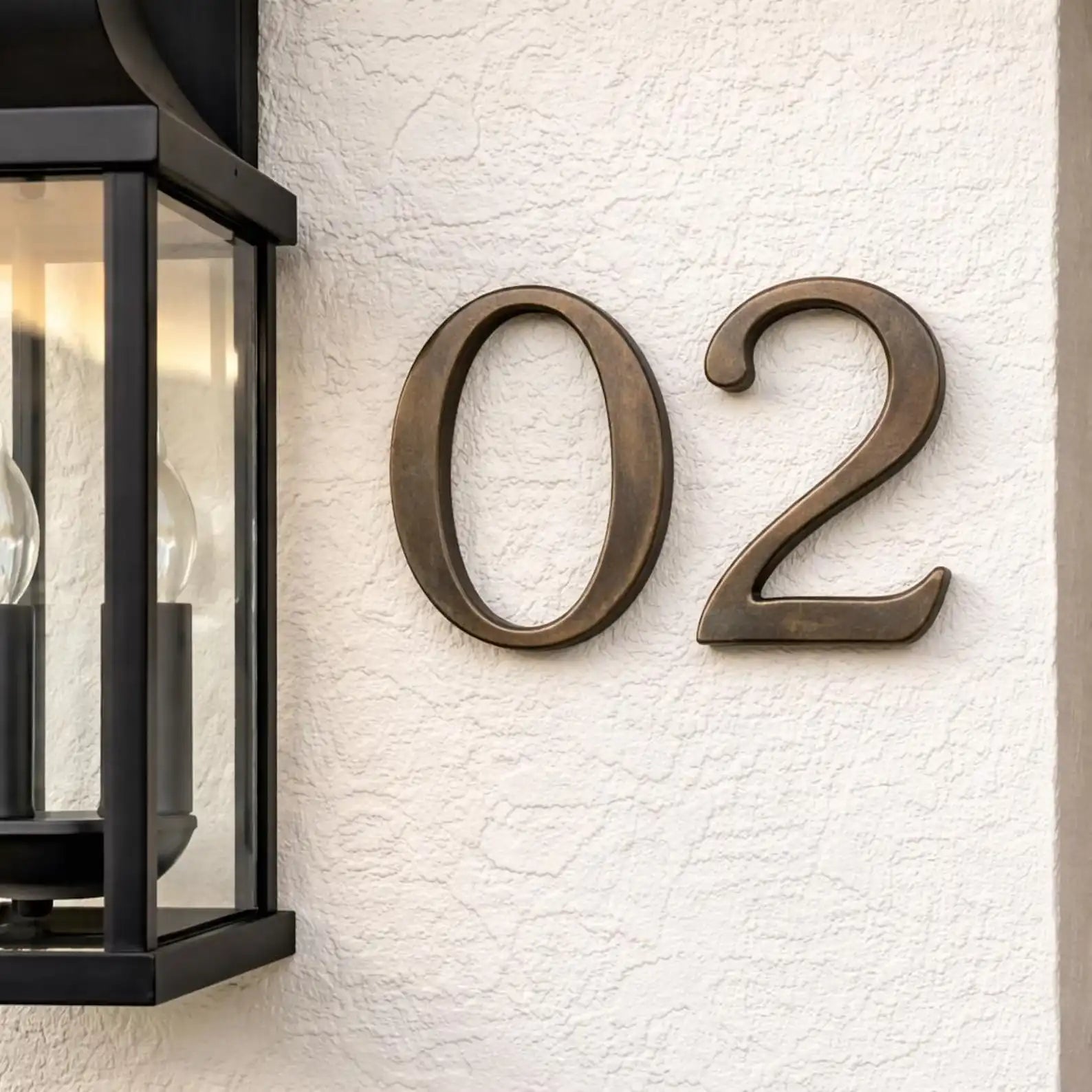 Bronze house numbers '02' on a textured white wall next to a black outdoor light fixture.