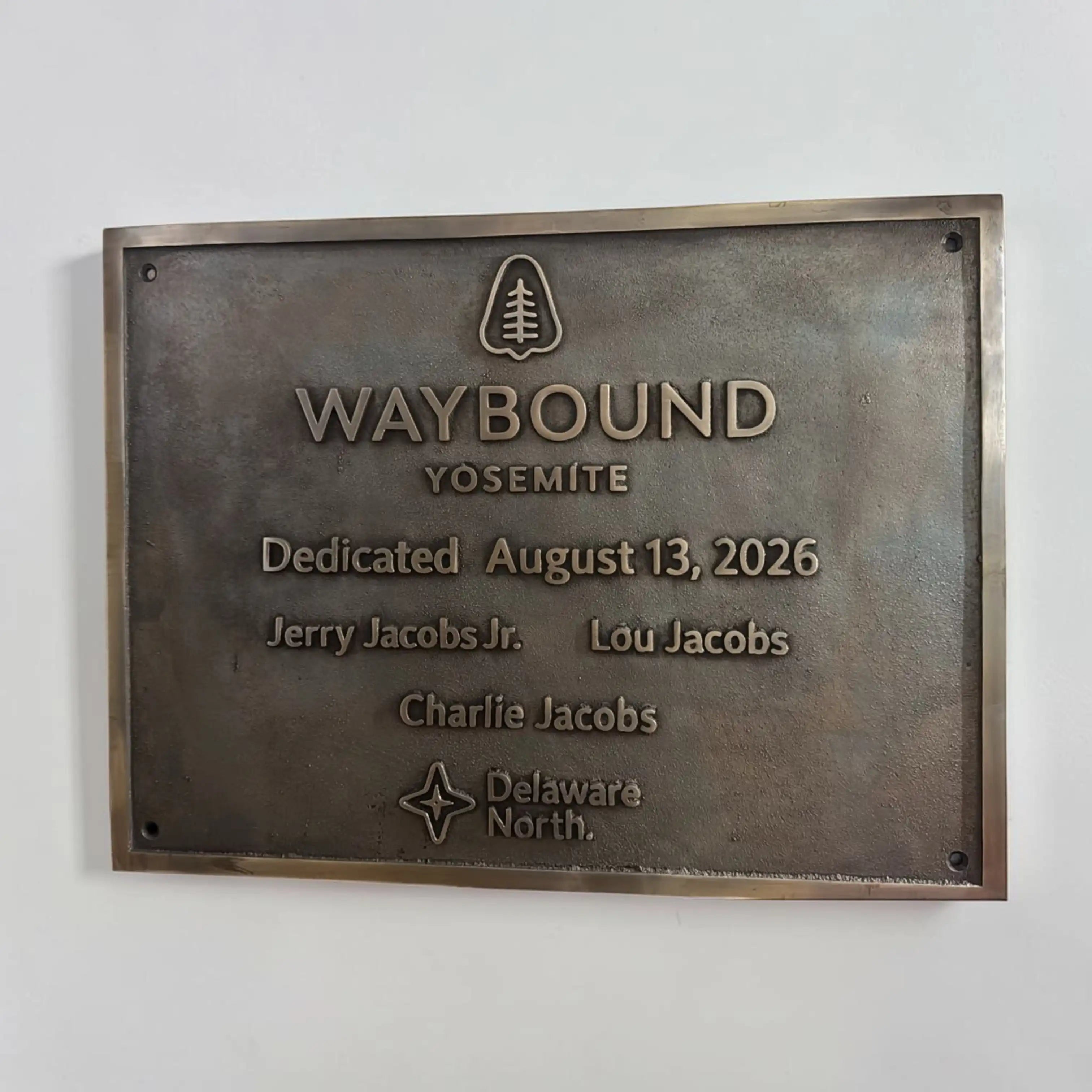 A rectangular bronze Waybound Yosemite memorial plaque mounted on a white wall. The plaque features raised text including 'WAYBOUND', 'Yosemite', the tree logo, the dedication date August 13, 2026, and names Jerry Jacobs Jr.