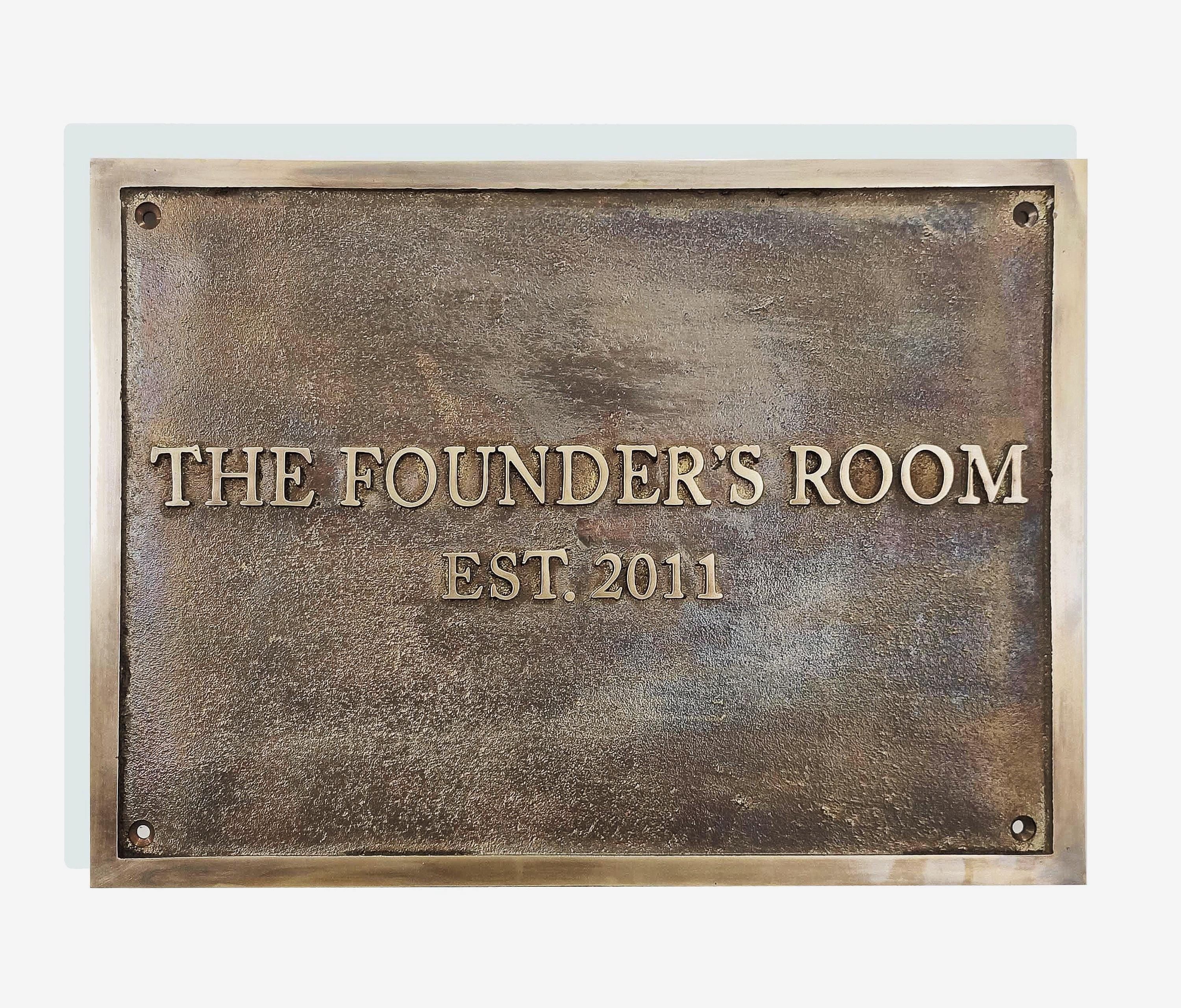 A cast bronze plaque with the inscription 'THE FOUNDER'S ROOM EST. 2011' in capital letters, set against a light background.