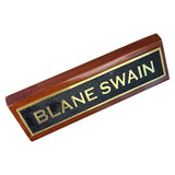 A rectangular wooden nameplate with a reddish-brown wood grain frame and a black textured center panel bordered in gold, featuring the name 'BLANE SWAIN' in gold lettering, shown on a plain white background.