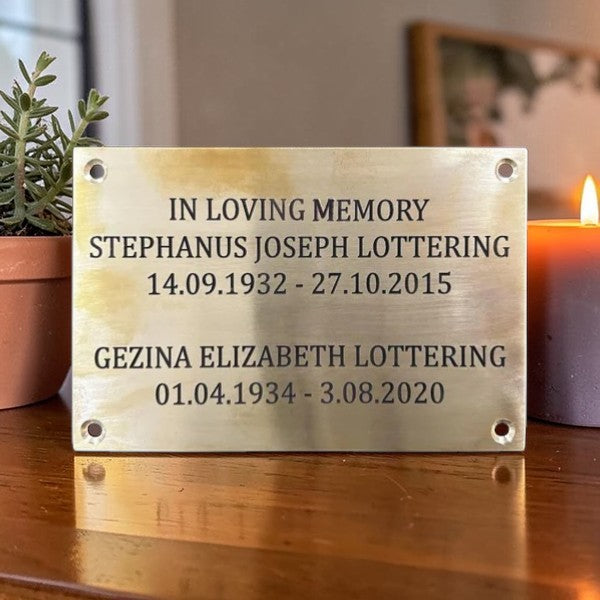Memorial plaque with names and dates on a wooden surface with a candle and plant in the background