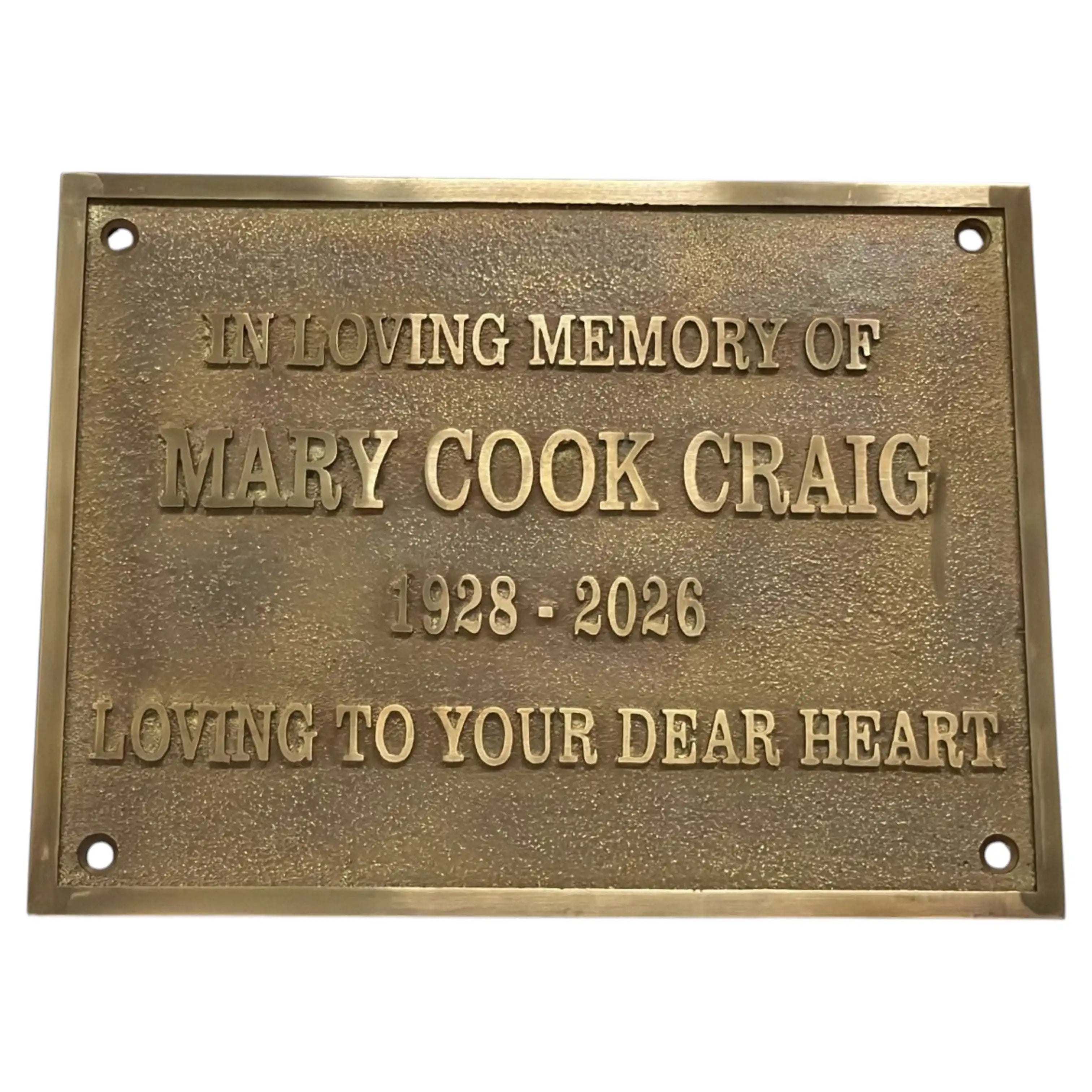 A rectangular bronze memorial plaque with raised lettering that reads 'IN LOVING MEMORY OF MARY COOK CRAIG 1928 - 2026 LOVING TO YOUR DEAR HEART' on a plain white background.